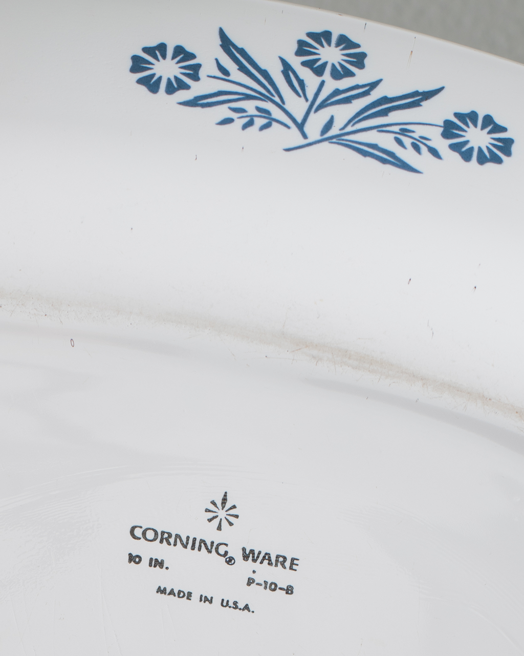 Close-up of the inside of a white Corning Ware plate or dish, showing a blue floral pattern near the top edge and printed Corning Ware branding near the bottom.