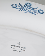 Close-up of the inside of a white Corning Ware plate or dish, showing a blue floral pattern near the top edge and printed Corning Ware branding near the bottom.