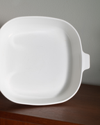 A white oval casserole dish decorated with a blue floral pattern, sitting on a wooden shelf against a light gray wall.