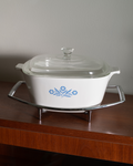 A white ceramic casserole dish with a glass lid and blue floral pattern, sitting on a silver metal trivet on a wooden table against a neutral wall.