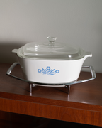 A white ceramic casserole dish with a glass lid and blue floral pattern, sitting on a silver metal trivet on a wooden table against a neutral wall.