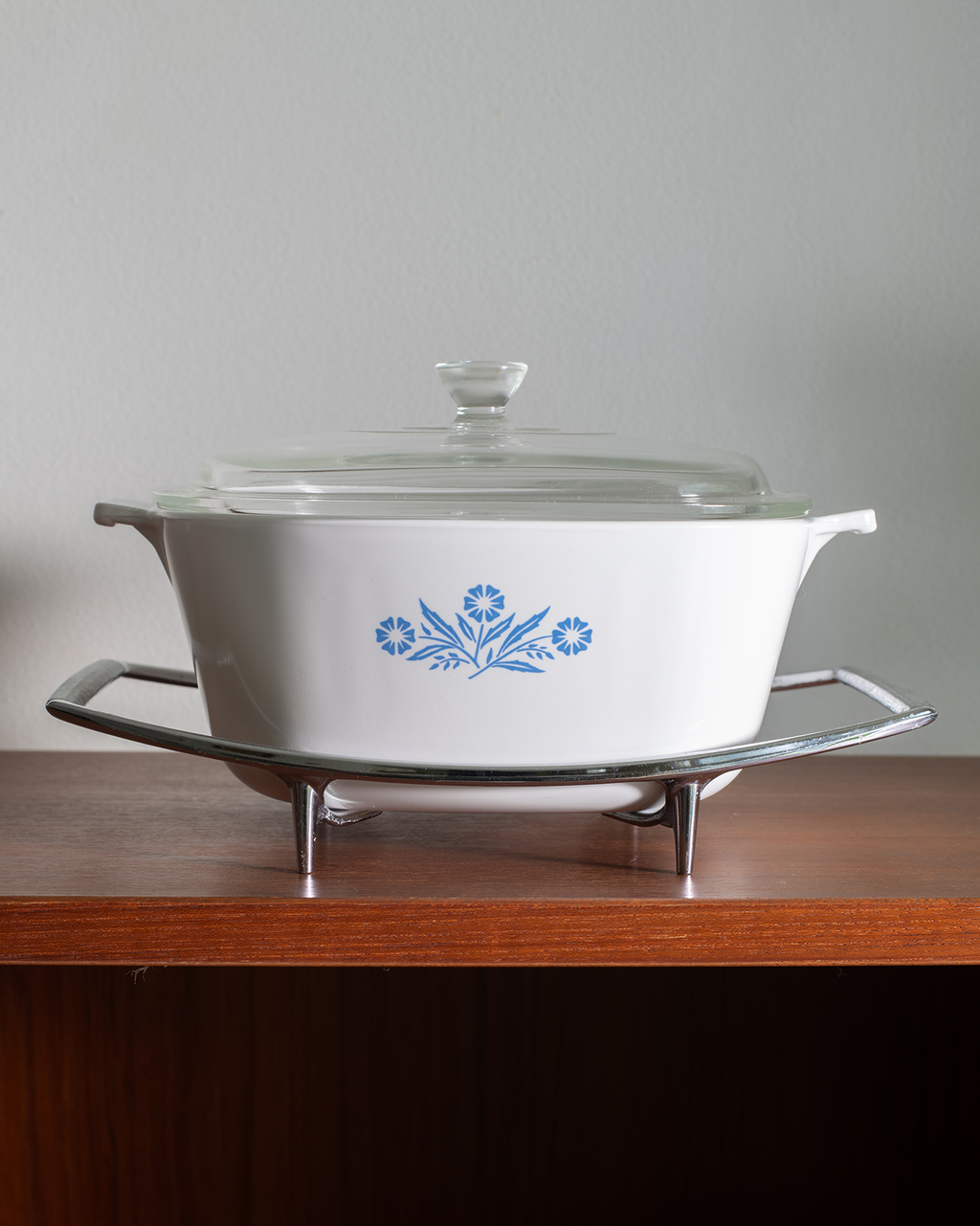 A white oval casserole dish with a glass lid, decorated with a blue floral pattern, sitting on a chrome stand on a wooden shelf against a light gray wall.