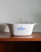 A white ceramic casserole dish with a glass lid, featuring a blue floral pattern on the front, sitting on a wooden shelf against a neutral gray wall.