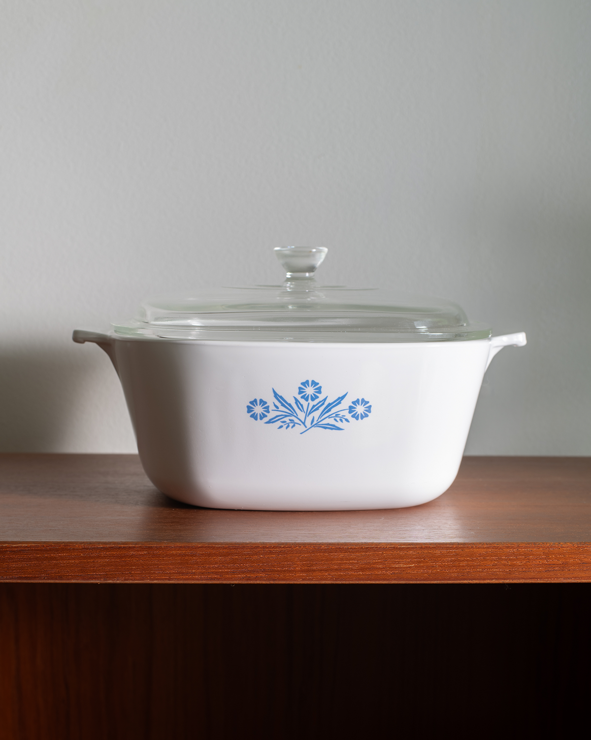 A white ceramic casserole dish with a glass lid, featuring a blue floral pattern on the front, sitting on a wooden shelf against a neutral gray wall.