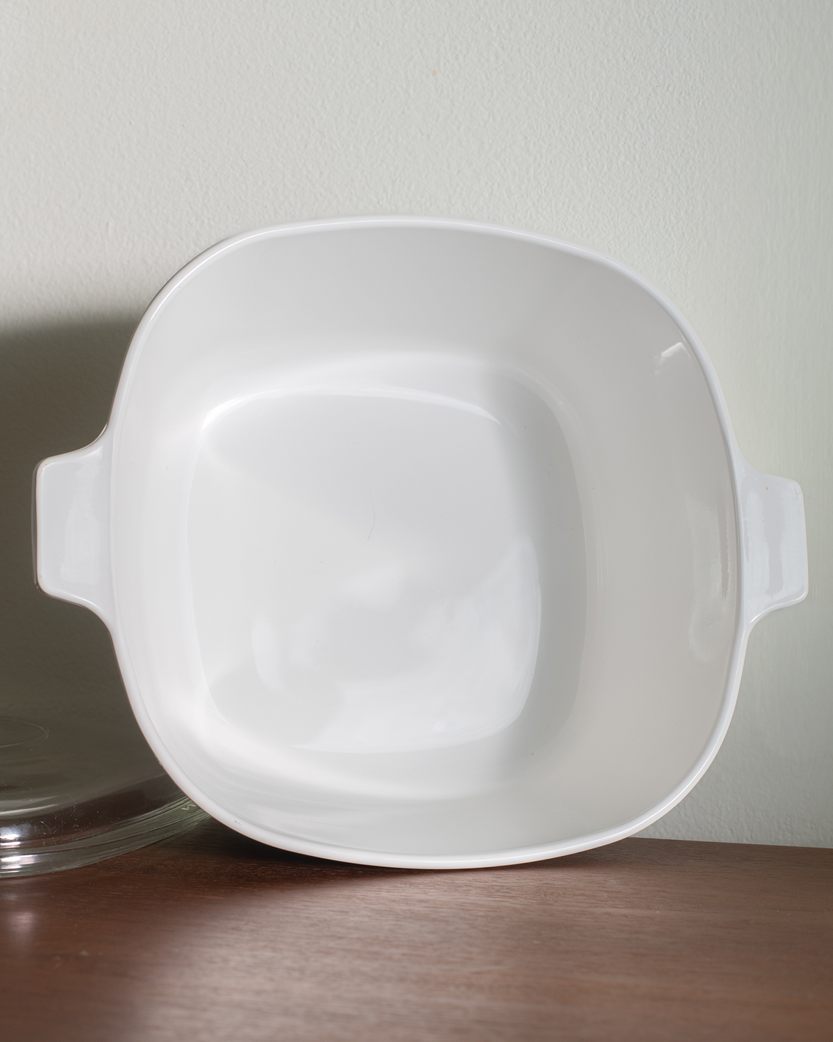 A white oval casserole dish with a glass lid, decorated with a blue floral pattern, sitting on a chrome stand on a wooden shelf against a light gray wall.
