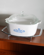 A white ceramic casserole dish with a clear glass lid and blue floral pattern, displayed on a metal stand on a wooden shelf against a light gray wall.