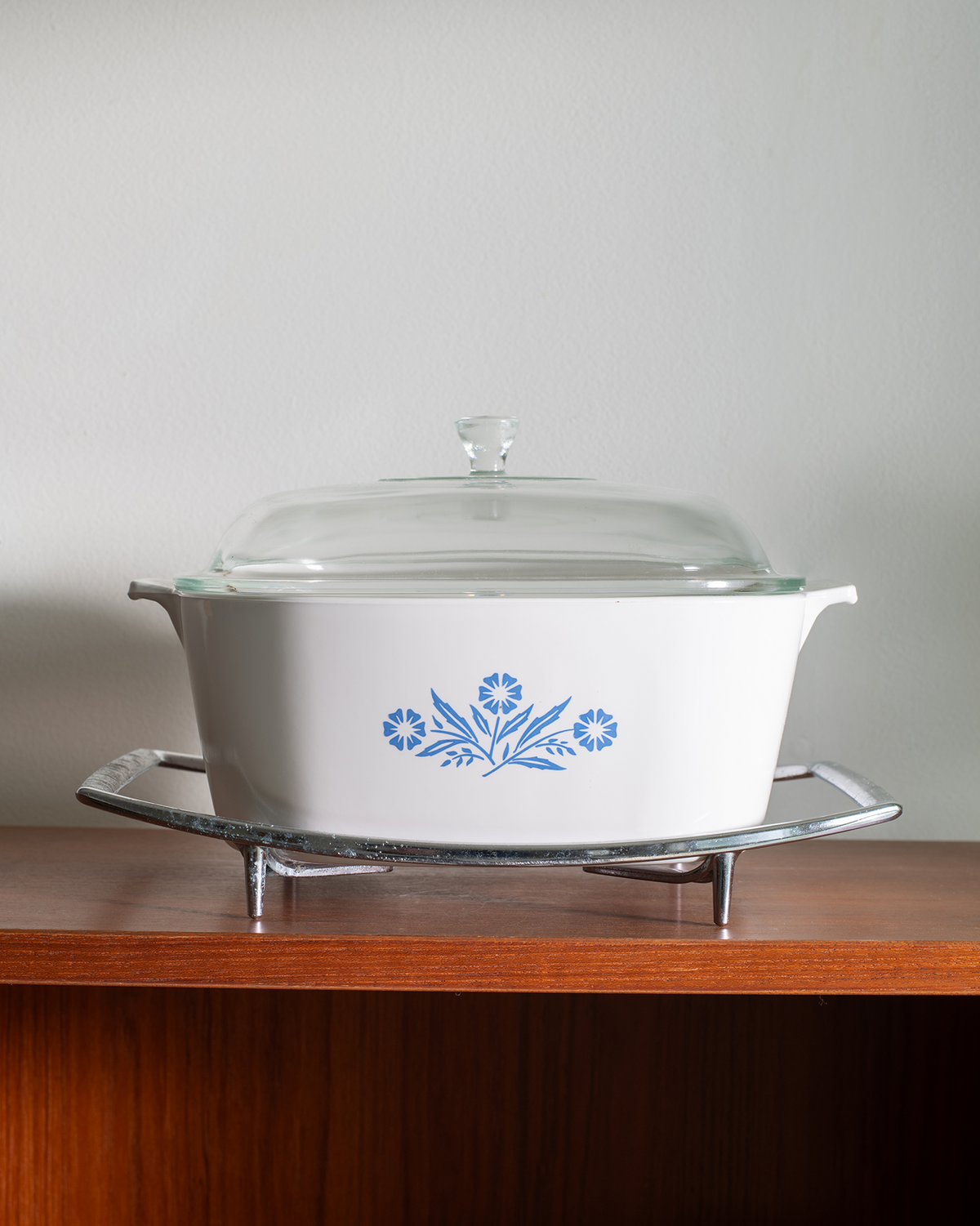 A white oval casserole dish with a blue floral pattern on the side, covered with a clear glass lid and sitting on a silver metal trivet, placed on a wooden shelf against a light gray wall.