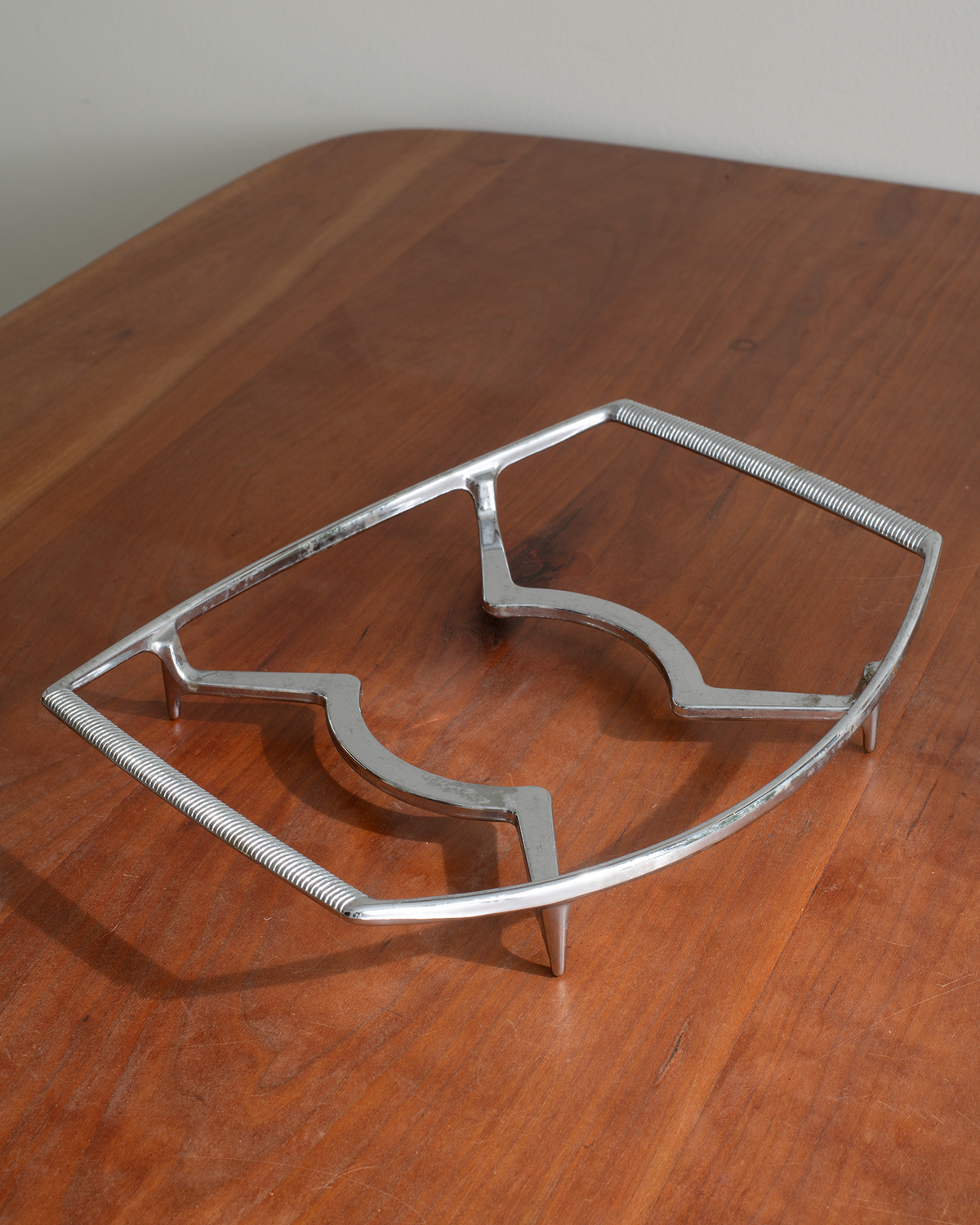 A silver metal trivet or support frame with a ribbed textured edge, displayed on a polished wooden table.