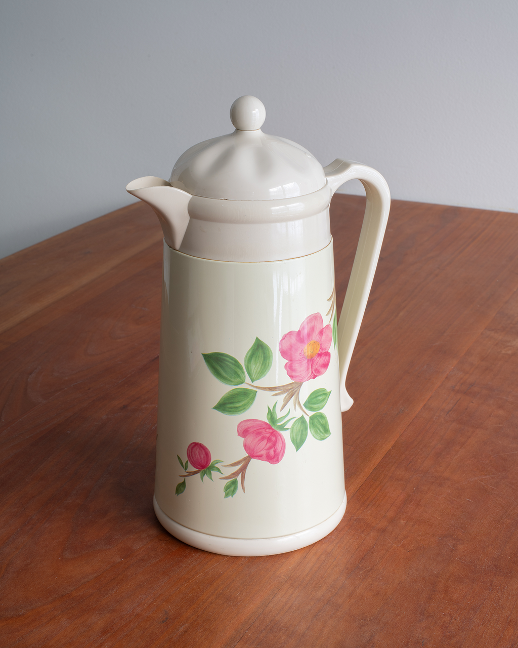 An off-white ceramic coffee carafe with a lid, featuring a pink rose and green leaf floral pattern on the front, set on a wooden table against a gray background.