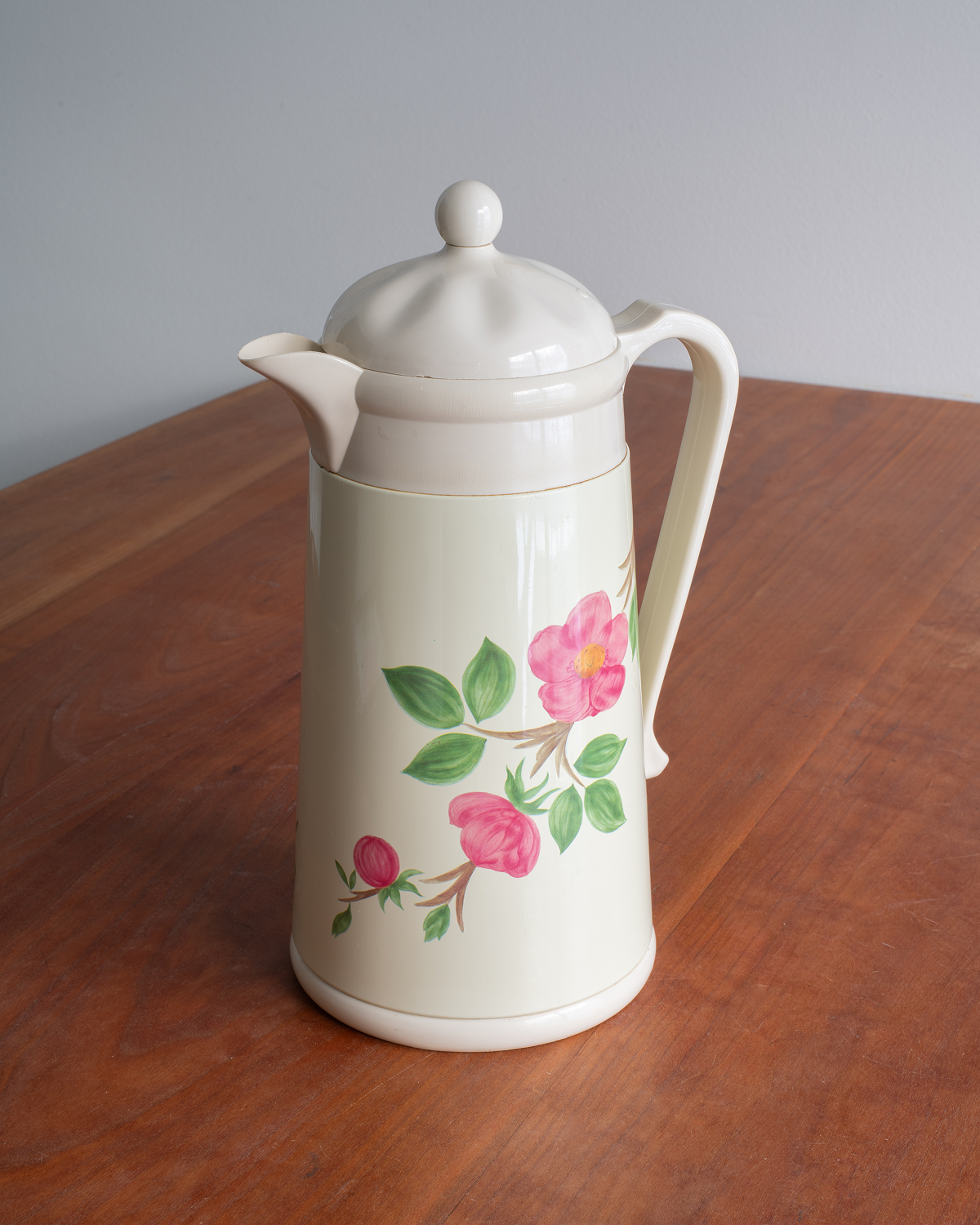 An off-white ceramic coffee carafe with a lid, featuring a pink rose and green leaf floral pattern on the front, set on a wooden table against a gray background.