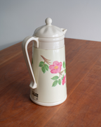An off-white vintage thermos coffee pot with a lid, featuring a pink rose and green leaves illustration on the front, sitting on a wooden table.