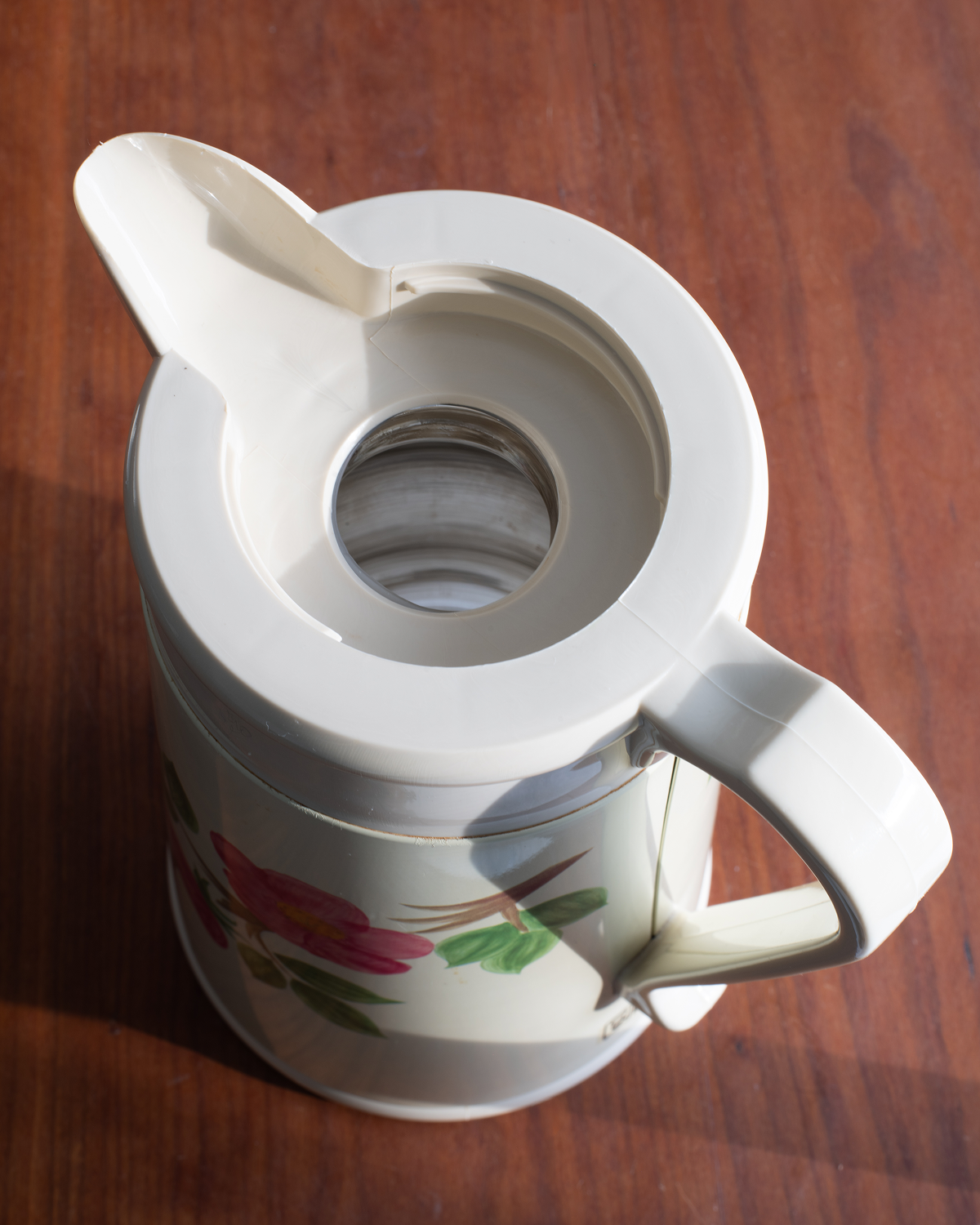 A white coffee pitcher with a floral print on the lower half, featuring pink flowers and green leaves, shown on a dark brown wooden surface.
