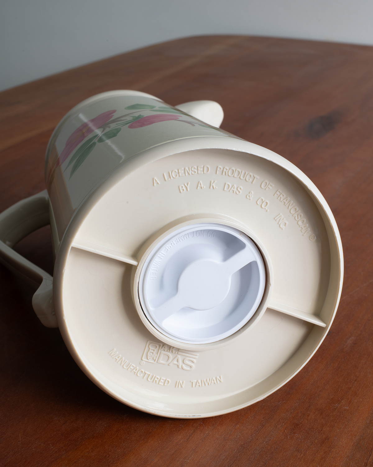 A vintage beige ceramic coffee thermos with a floral pattern, shown from a top-down angle on a wooden tabletop. The lid has a white plastic pour-over spout.