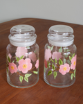 Two clear glass canisters with clear lids, decorated with pink flowers and green leaves, sitting on a wooden table against a neutral background.