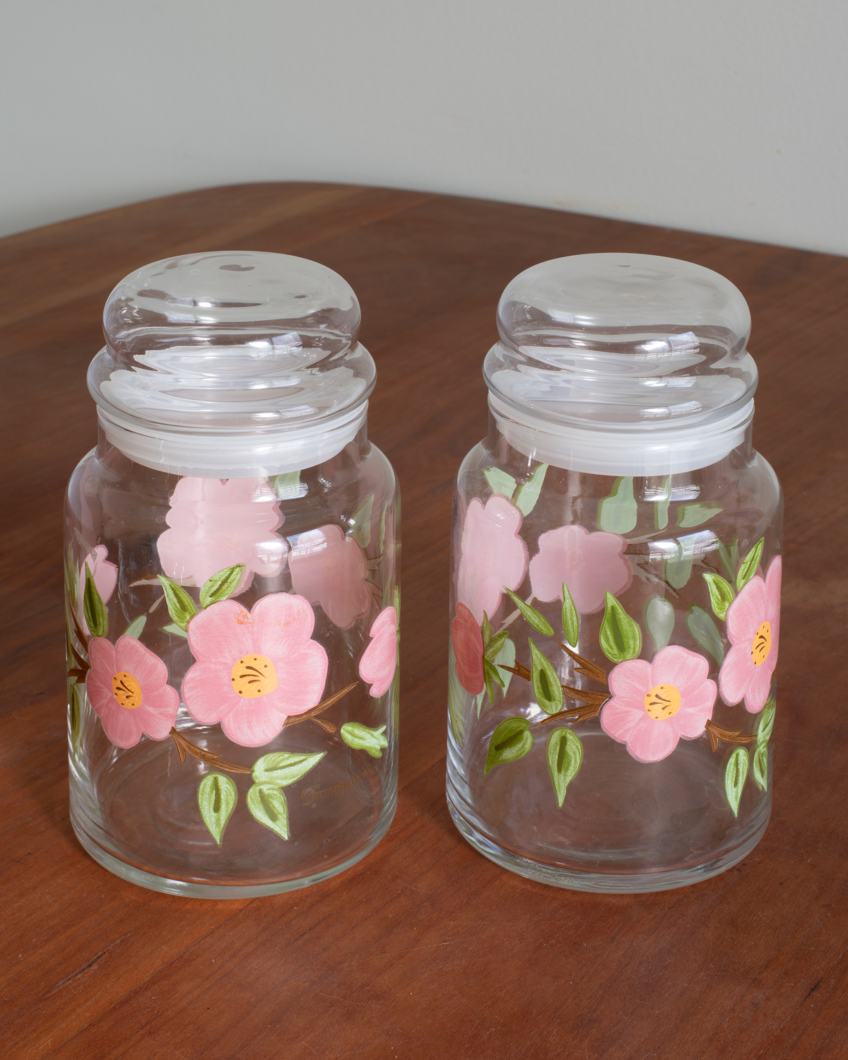 Two clear glass canisters with clear lids, decorated with pink flowers and green leaves, sitting on a wooden table against a neutral background.
