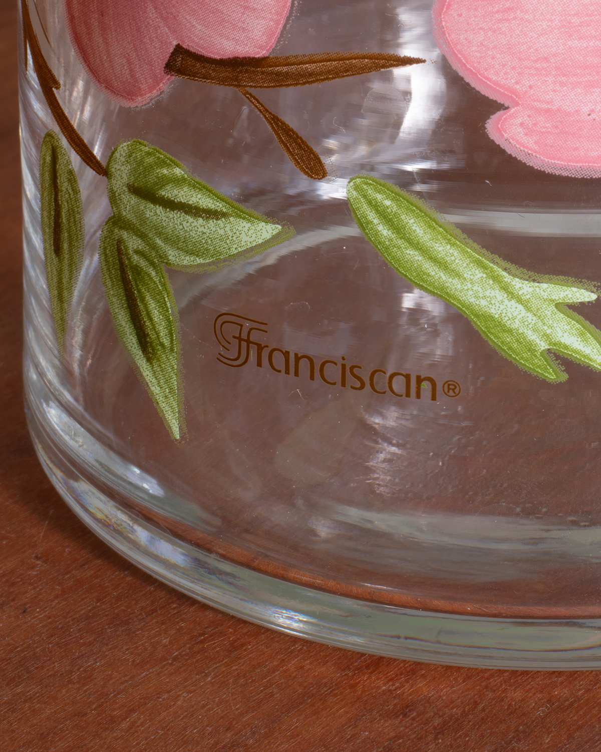 Close-up of a clear glass container or vase with pink floral decoration and green leaves, featuring the brand name 'Franciscan' printed in brown on the side, resting on a wooden surface.