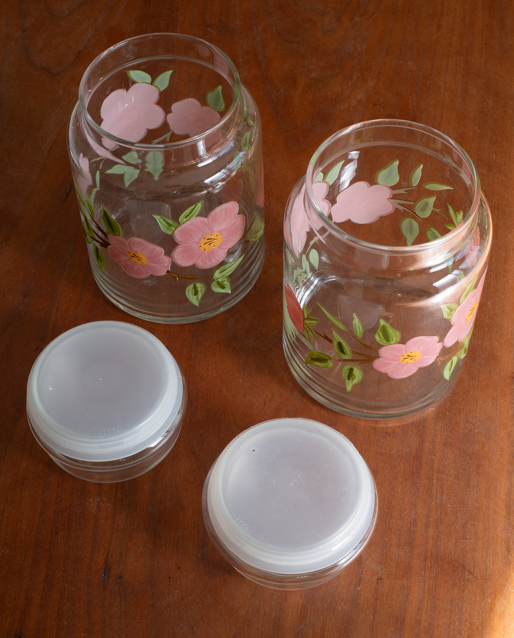 Two clear glass jars with pink cherry blossom flower and green leaf hand-painted designs, shown on a wooden table alongside two white plastic lids.