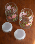 Two clear glass jars with pink cherry blossom flower and green leaf hand-painted designs, shown on a wooden table alongside two white plastic lids.