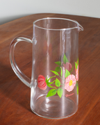 A clear glass pitcher with a floral print of pink flowers and green leaves, sitting on a wooden table.