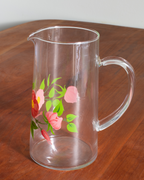 A clear glass pitcher with a floral print of pink and red flowers and green leaves, sitting on a wooden table.