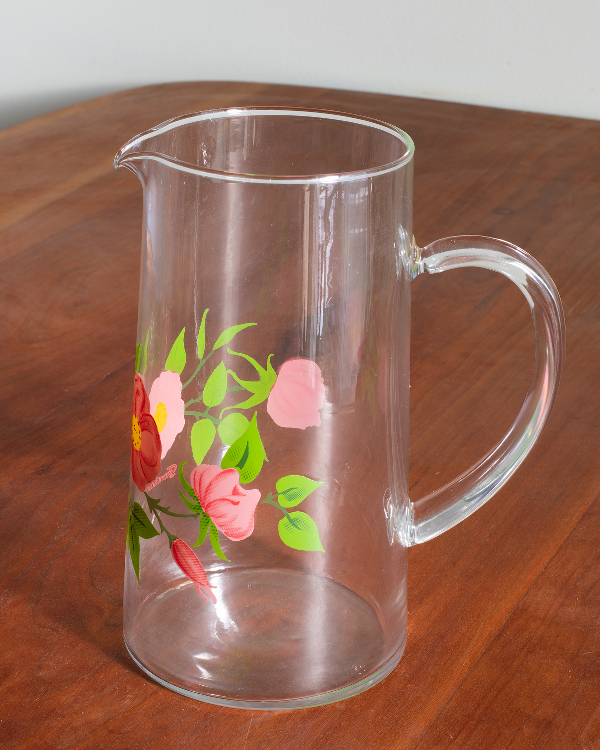 A clear glass pitcher with a floral print of pink and red flowers and green leaves, sitting on a wooden table.