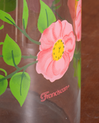 A close-up of a clear glass bottle featuring a pink rose with a yellow center and green leaves, printed with the brand name 'Franciscan' in red near the bottom.