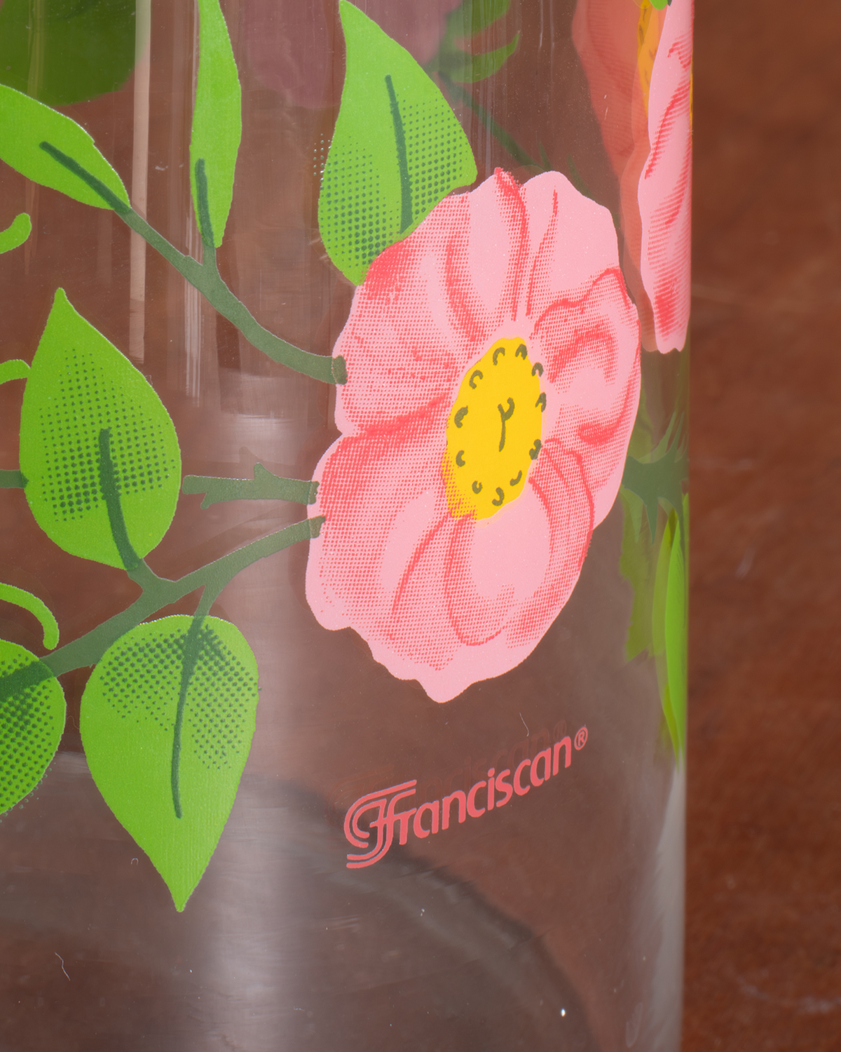 A close-up of a clear glass bottle featuring a pink rose with a yellow center and green leaves, printed with the brand name 'Franciscan' in red near the bottom.
