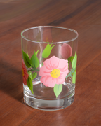 A clear glass tumbler with a thick base, decorated with a hand-painted pink flower, green leaves, and red accents, set on a dark wooden table.