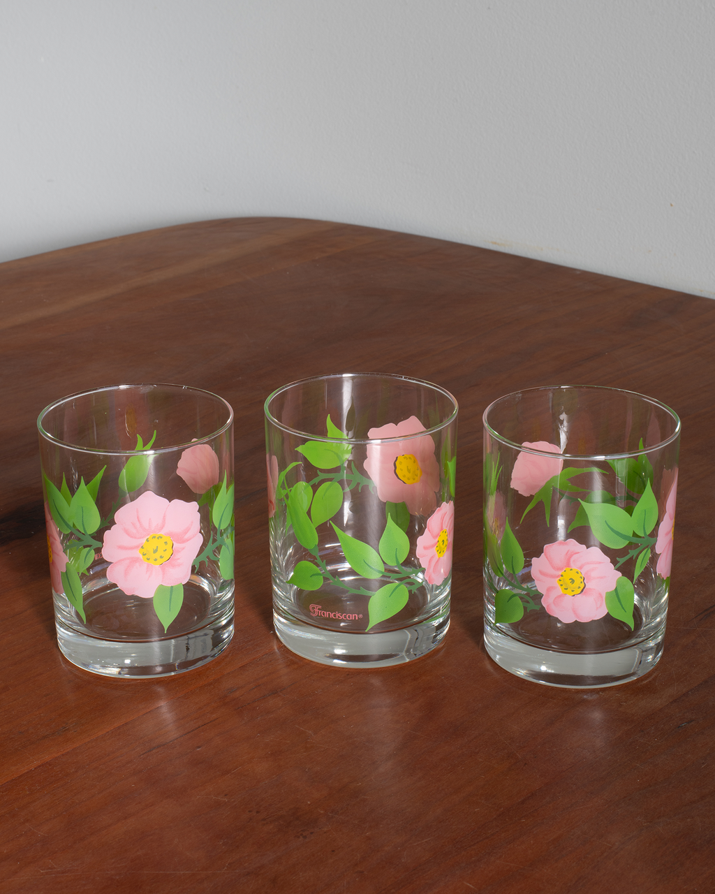 Three clear glass tumblers with pink flowers and green leaves printed on them, arranged on a wooden tabletop against a light gray wall.