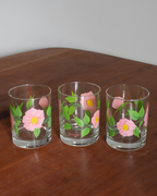 Three clear glass tumblers with pink flowers and green leaves printed on them, arranged on a wooden tabletop against a light gray wall.