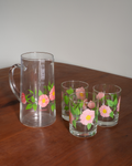 A clear glass pitcher and two matching drinking glasses with pink floral prints and green leaves, arranged on a wooden table against a plain light wall.
