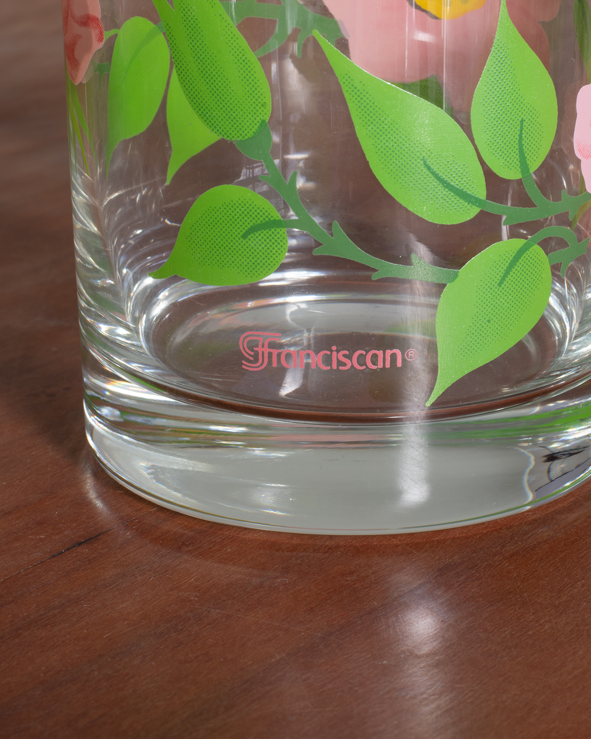 Close-up of a clear glass tumbler with a green leaf and pink flower pattern, resting on a dark wooden table. The brand name 'Franciscan' is printed in pink on the base.