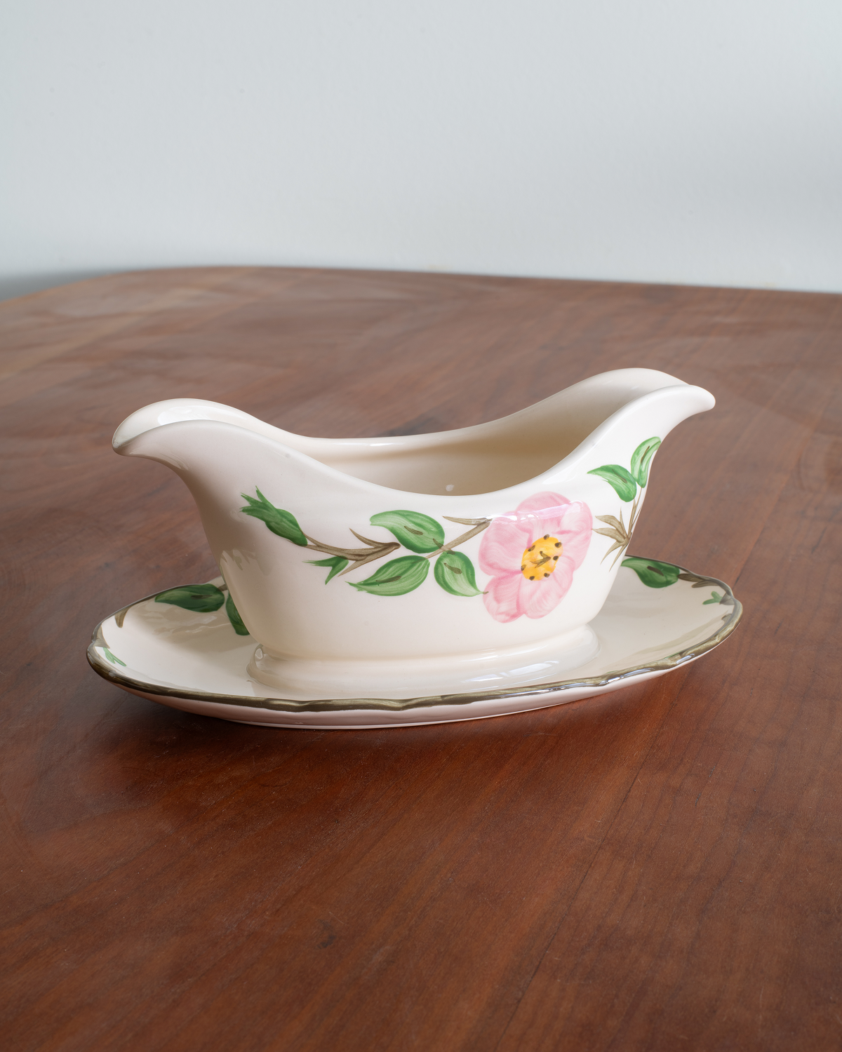 A vintage cream-colored porcelain gravy boat with a matching saucer, decorated with a pink rose and green leaves, resting on a wooden table.