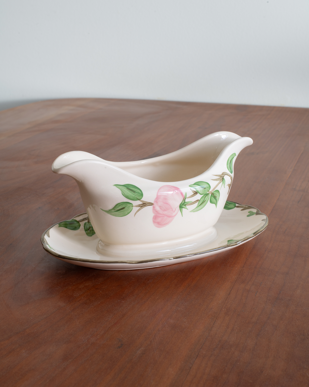 A vintage ceramic gravy boat with a matching saucer, decorated with a pink rose and green leaves on an off-white background, set on a wooden table.