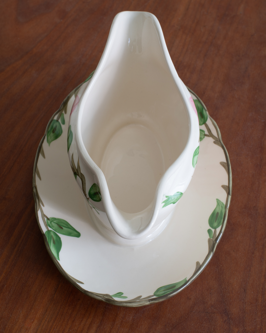 A white porcelain gravy boat with a matching saucer, decorated with a green leaf and brown vine pattern, set on a dark wooden table.