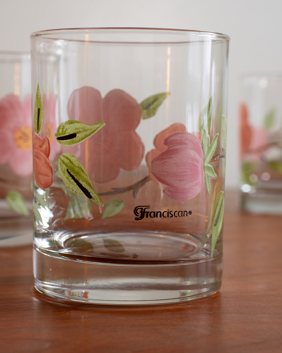 A clear glass tumbler with hand-painted pink flowers and green leaves, featuring the brand name 'Franciscan' printed in black script near the bottom, set on a wooden table.