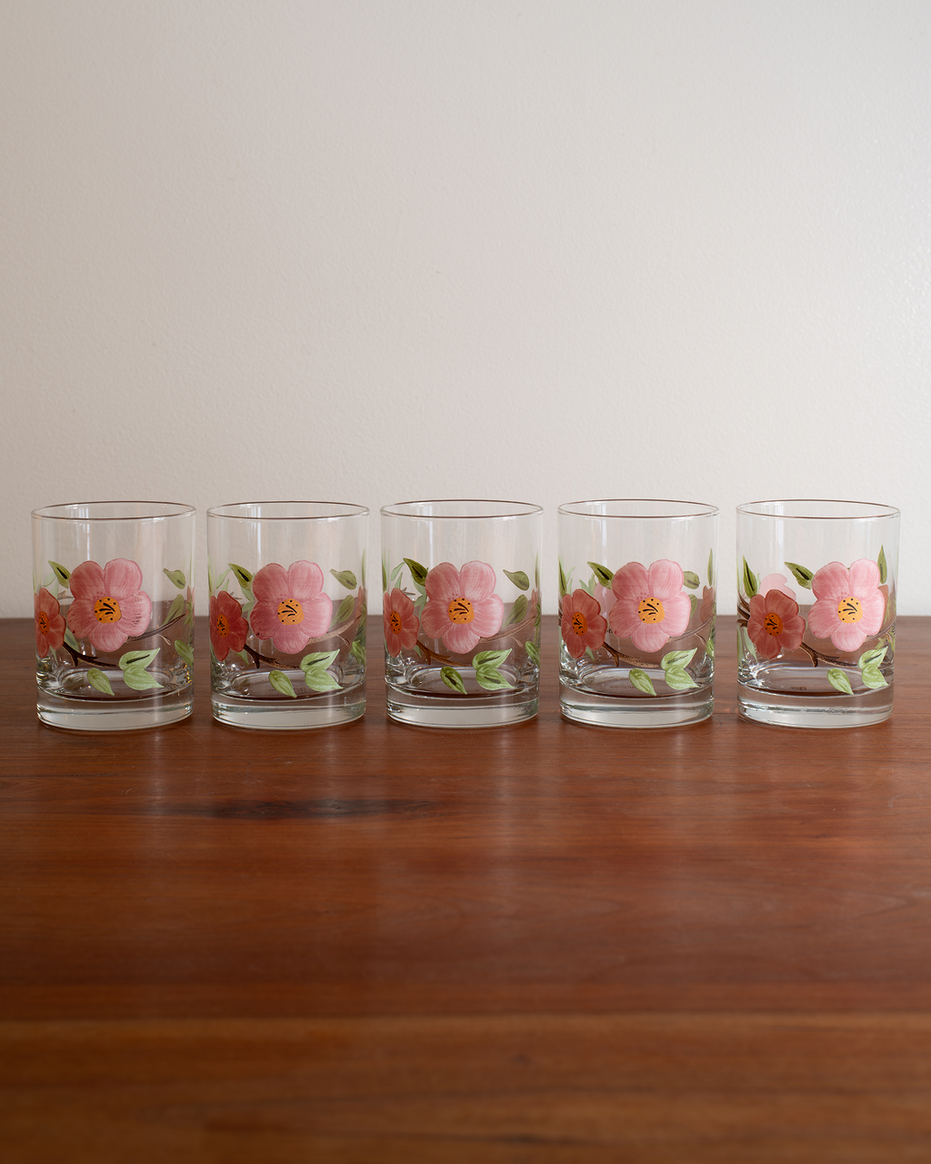 Five clear glass tumblers with pink and orange floral designs, arranged in a row on a wooden table against a plain light background.