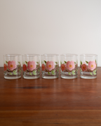 Five clear glass tumblers with pink and orange floral designs, arranged in a row on a wooden table against a plain light background.