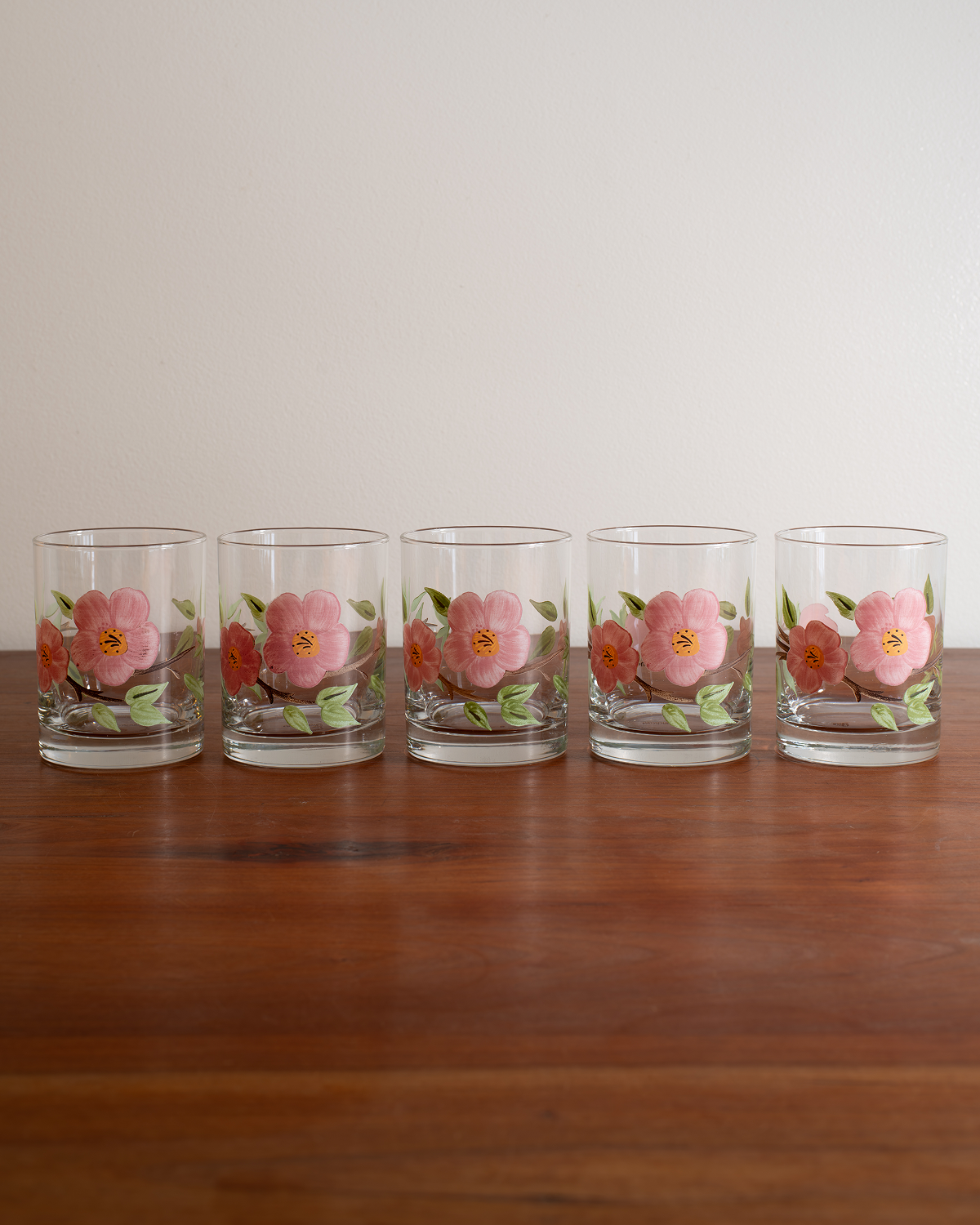 Five clear glass tumblers with pink and orange floral designs, arranged in a row on a wooden table against a plain light background.