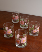 Four clear glass tumblers with hand-painted pink flowers and green leaves, arranged on a dark wooden table against a plain light wall.