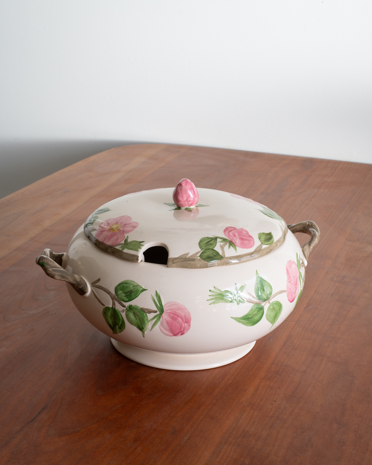 A ceramic lidded serving caddy or tureen with pink rose motifs and green leaves, placed on a wooden table against a plain light background.