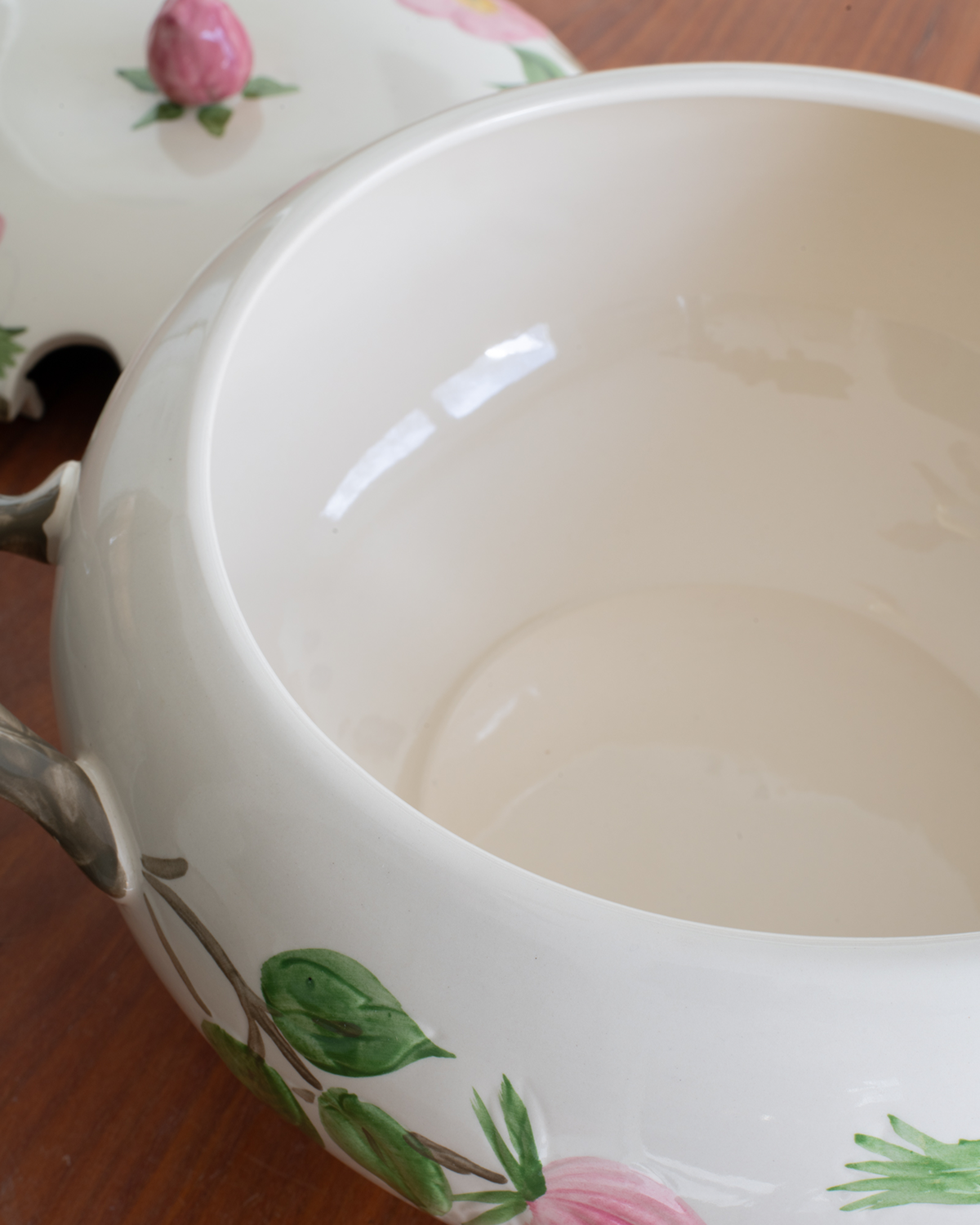 Close-up of a white soup toureen with a pink rose and green leaf floral pattern, photographed on a wooden surface.