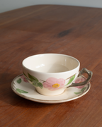 A cream-colored porcelain teacup and saucer set with a pink flower and green leaf pattern, resting on a wooden table.