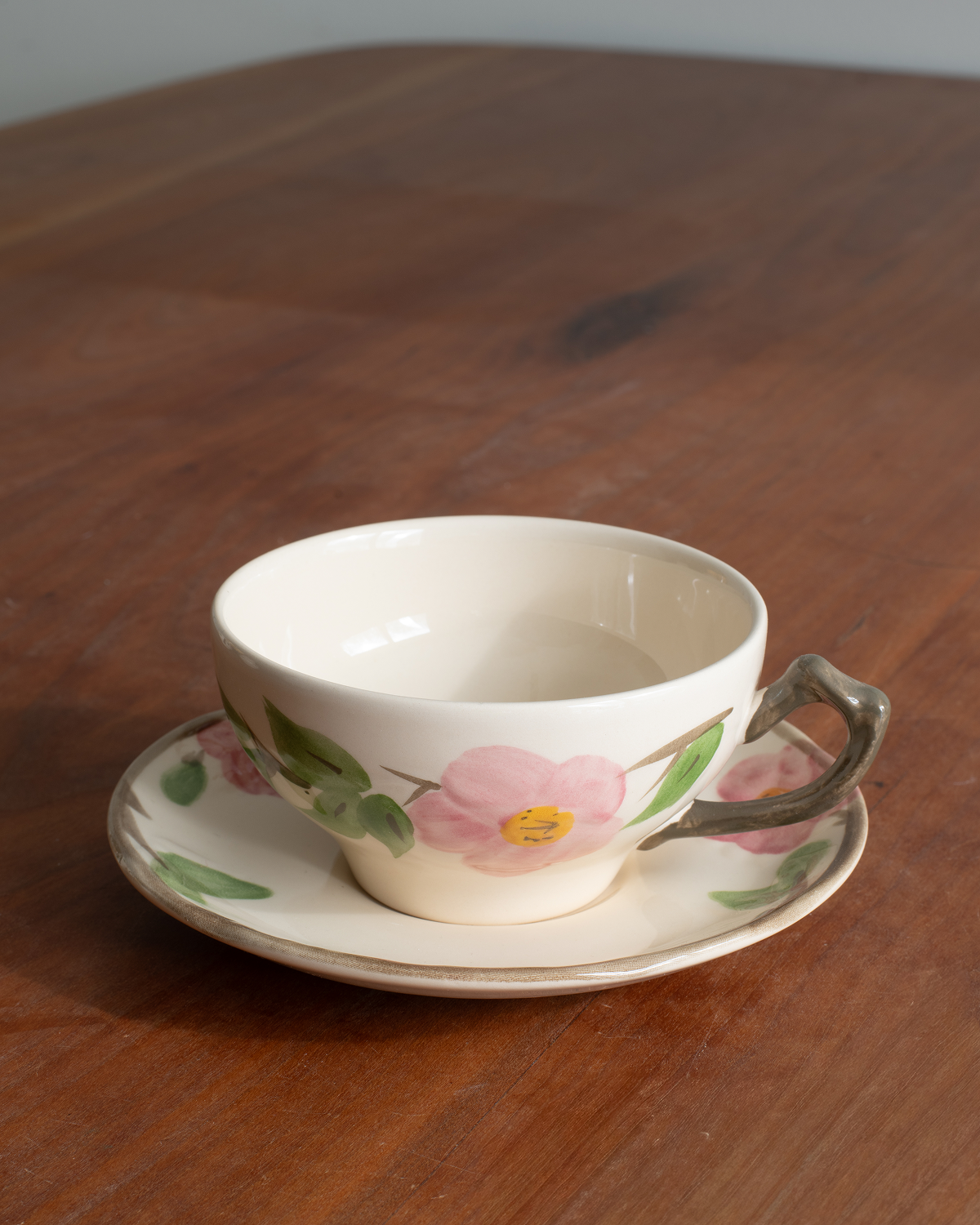 A cream-colored porcelain teacup and saucer set with a pink flower and green leaf pattern, resting on a wooden table.