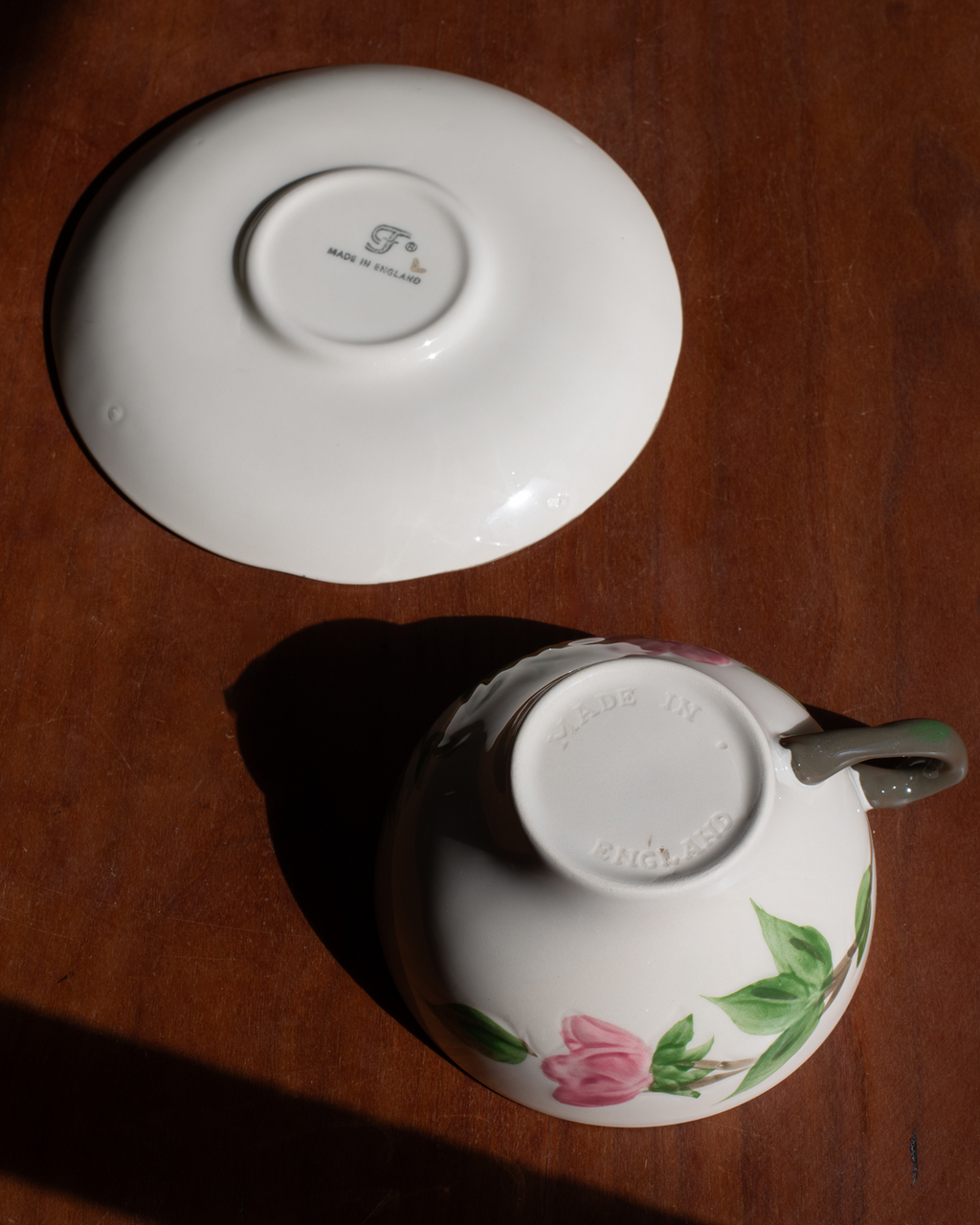 A white porcelain teacup and saucer set on a dark wooden table, photographed from above in direct sunlight. The saucer shows a printed logo in the center, and the cup is decorated with a pink flower and green leaves near the base.