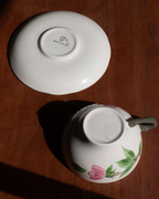 A white porcelain teacup and saucer set on a dark wooden table, photographed from above in direct sunlight. The saucer shows a printed logo in the center, and the cup is decorated with a pink flower and green leaves near the base.