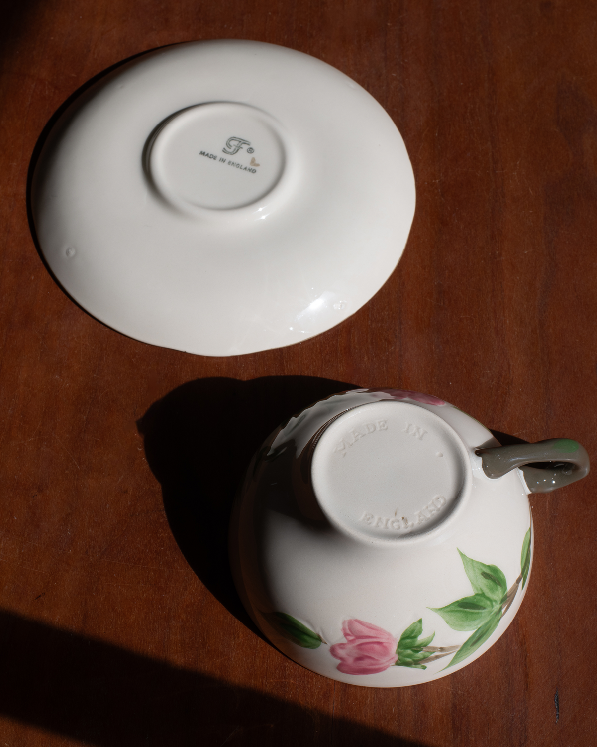 A white porcelain teacup and saucer set on a dark wooden table, photographed from above in direct sunlight. The saucer shows a printed logo in the center, and the cup is decorated with a pink flower and green leaves near the base.