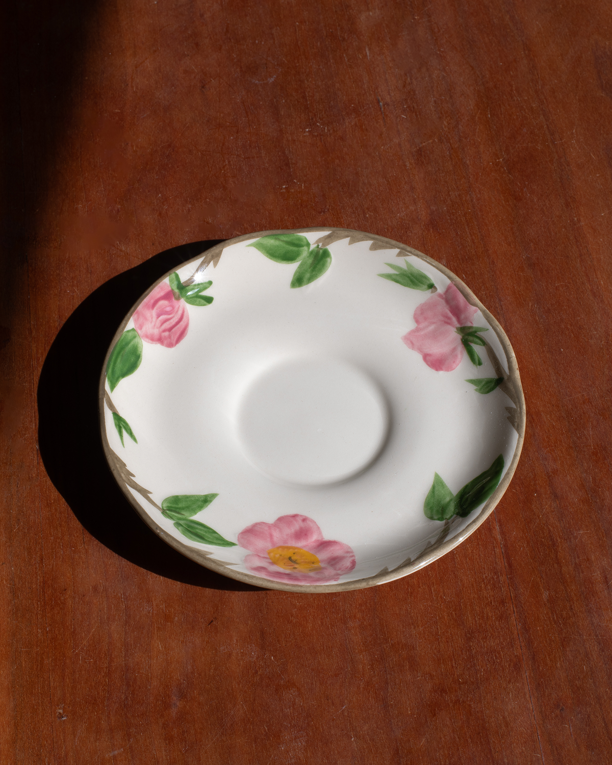 A white ceramic saucer decorated with pink flowers and green leaves, placed on a warm brown wooden surface under direct sunlight.