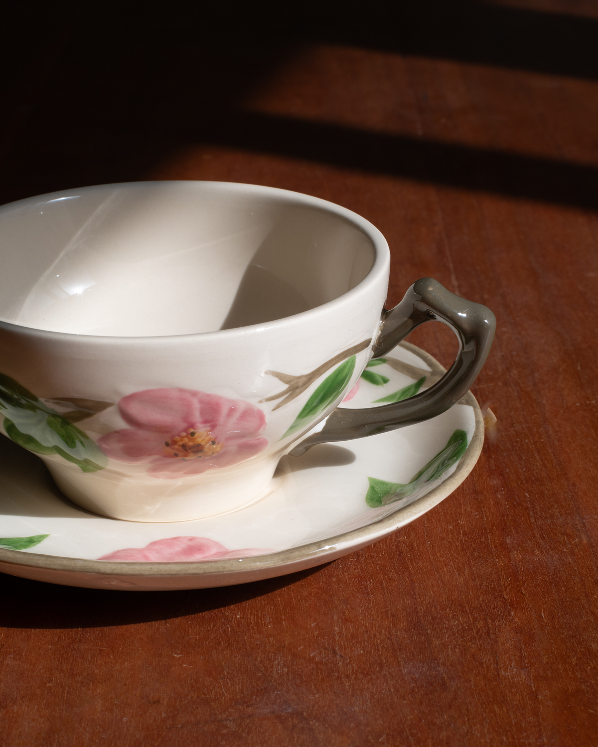 A white ceramic teacup and saucer set with a pink floral print and green leaves, featuring a dark gray handle, resting on a dark wooden table in warm sunlight.