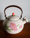 A cream-colored enamel tea kettle with a pink floral design on the side, featuring a dark brown handle and lid topper, resting on a wooden surface against a plain light gray wall.
